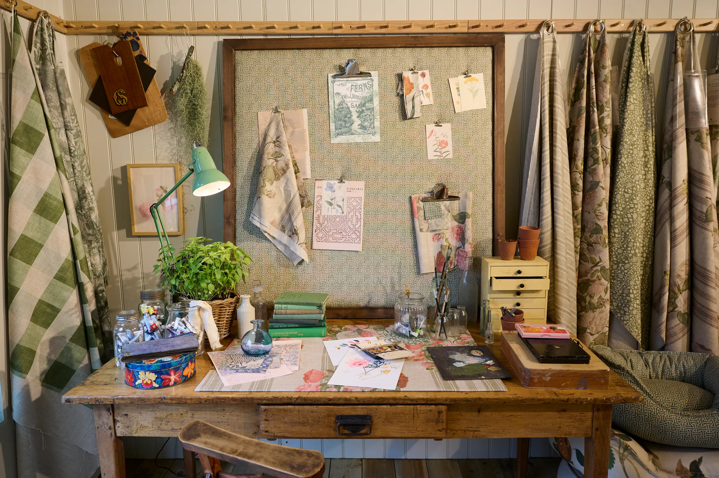 Recreated artist’s studio at the RHS Chelsea Flower Show, featuring a wooden worktable with botanical sketches, paints and books, fabric swatches pinned to a board, and floral textiles hanging around the space.